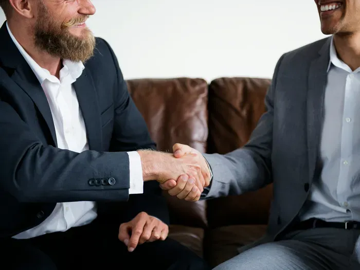 Two smiling businessmen in suits shaking hands confidently while seated on a leather couch in office setting